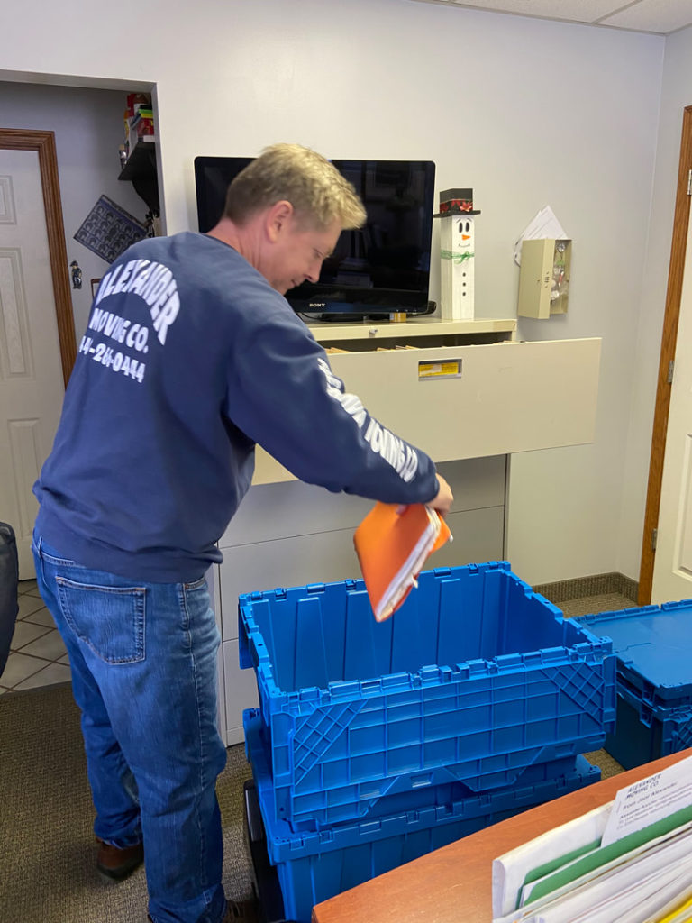 Man in blue shirt packing folders into blue crates. TV, file cabinet, and snowman decor visible in office.